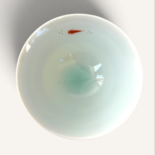 A top-down view of a white ceramic bowl with a glossy finish, featuring a small orange fish illustration near the top edge and a soft light blue-green reflection in the center, set against a plain light gray background.