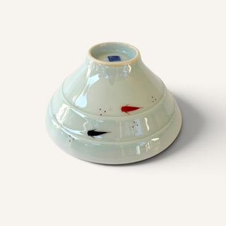 A glossy ceramic bowl with a light green glaze, featuring hand-painted abstract black and red brushstrokes on the exterior, photographed on a clean off-white background.