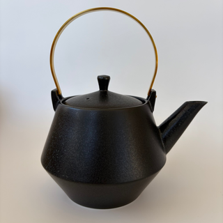 A matte black ceramic teapot with a textured glaze, featuring a gold-colored metal loop handle and a matching gold spout accent.
