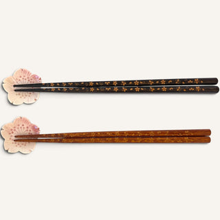 Two pairs of chopsticks displayed horizontally on a light cream background, each resting on a small pink flower-shaped stand. The top pair is black with small gold floral patterns, and the bottom pair is a warm brown wood tone with matching gold floral patterns.