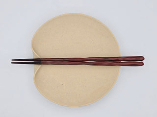 Toki Meki Workshop red lacquered wooden chopsticks with a wavy design, resting on a speckled beige plate.